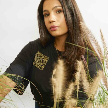 Woman sitting amongst grasses wearing the rose embroidery in a square on the upper right of the black Asmuss Evelyne Tshirt that has been hand embellished by Asmuss designer