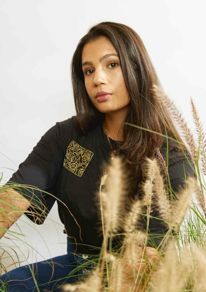 Woman sitting amongst grasses wearing the rose embroidery in a square on the upper right of the black Asmuss Evelyne Tshirt that has been hand embellished by Asmuss designer