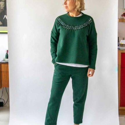 Woman, with short wavy blond hair, wearing the Asmuss Curve Joggers in Trekking Green and the Anni Sweatshirt in Trekking Green, standing with weight on one foot and looking to her right