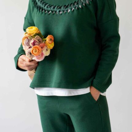 1.	Close up of a woman wearing the Asmuss Anni Sweatshirt in Trekking Green with the Cut Twist and Stitch detailing while holding a bunch of yellow, orange and pink flowers under her arm