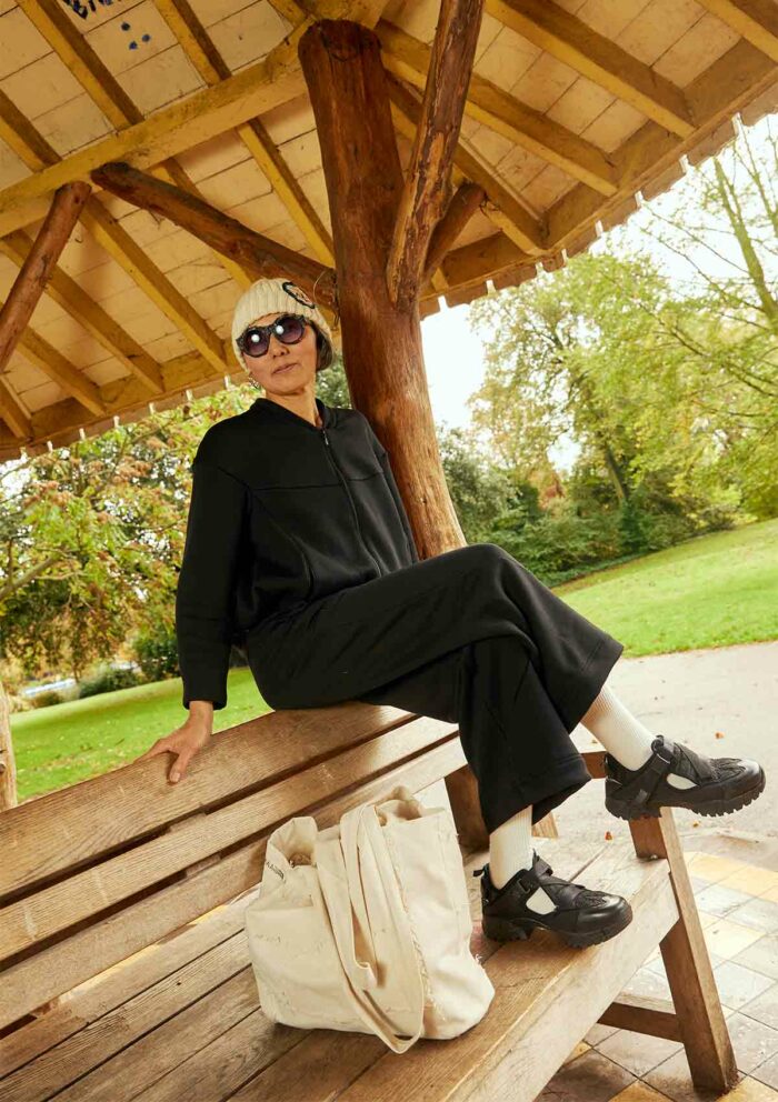 Woman sitting on bench in gazebo, wearing the Asmuss Hepworth zip through sweat, Column joggers in black and also an Asmuss hand knitted ecru beanie. The Asmuss Scraps Tote bag is in the foreground sitting on the bench seat beside the women.