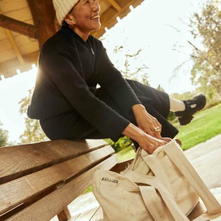 Woman sitting on bench in gazebo, wearing the Asmuss Hepworth zip through sweat, Column joggers in black and also an Asmuss hand knitted ecru beanie. The women is laughing and reaching down into the Asmuss Scraps Tote bag