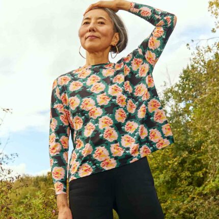 Woman wearing the Asmuss Paige Long Sleeve T-shirt in geometric rose print.  She has one hand on her head and is standing in front of trees