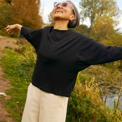 Woman looking up into the sky with her arms out.  You can see the bat wing sleeves of the Asmuss Puzzle Top that she is wearing.  She is standing in a field with trees and pond behind her.