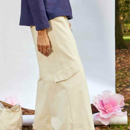 Woman standing side on wearing the Asmuss Eddy Cargo Trousers and Asmuss Anni Sweatshirt in Navy. The hem buttons are loose. She is standing in front of a white background, weighted down by a branch in a park with an Asmuss Tote Bag and pink paper flower on the ground behind her