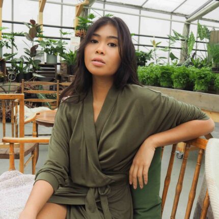 A woman wearing the AWAN Wrap Dress in Olive Green, sitting in a greenhouse surrounded by lush plants. She poses elegantly on a wooden chair, showcasing the dress's relaxed fit and adjustable waist belt.