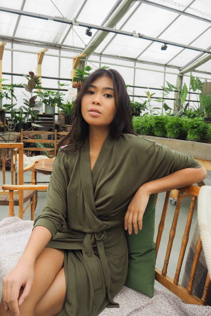 A woman wearing the AWAN Wrap Dress in Olive Green, sitting in a greenhouse surrounded by lush plants. She poses elegantly on a wooden chair, showcasing the dress's relaxed fit and adjustable waist belt.