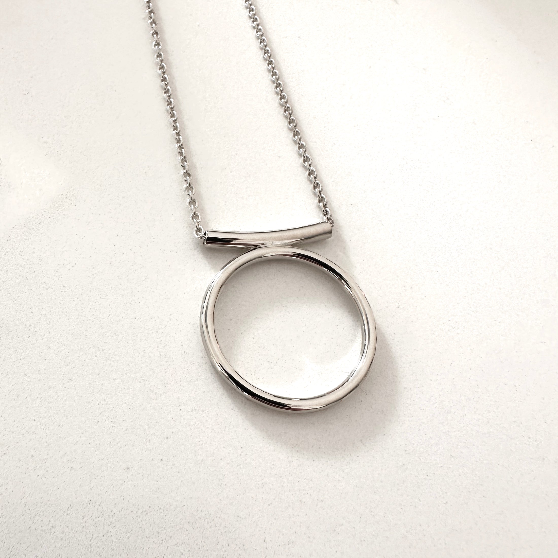 A silver-colored circle necklace with a rolo-style chain and a pendant featuring a circle with a curved line above it.