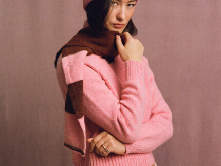 Model in three-quarter view wearing a pink ribbed beanie with blue trim, styled with a pink sweater and scarf. Made in Britain.